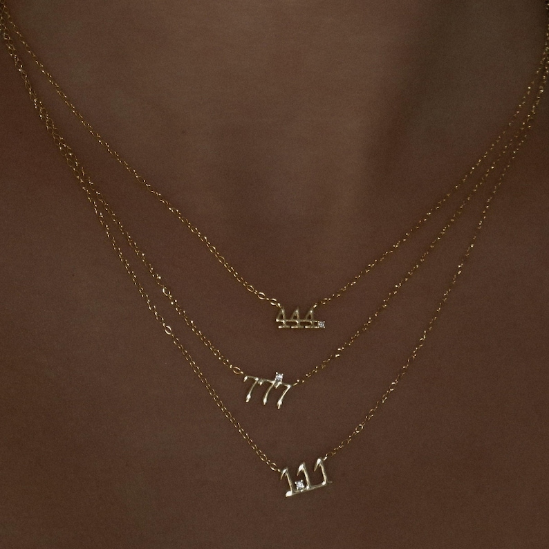 May include: Three gold necklaces with pendant numbers 111, 777, and 444. Each necklace has a fine chain. The pendants are in a script font and have small, clear gemstones. The necklaces are layered and worn on a person.
