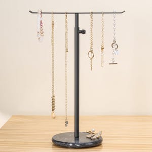 May include: A black metal T-shaped jewelry stand with an adjustable height pole and a round, dark gray marble base. The stand holds several necklaces and bracelets, including beaded, gold-tone chain, and pearl-style jewelry pieces.
