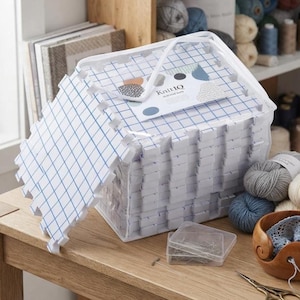 May include: A stack of white interlocking foam blocking mats with a blue grid pattern, housed in a clear plastic zippered storage bag labeled KnitIQ. The set includes a small clear container of T-pins, yarn balls, and metal scissors.
