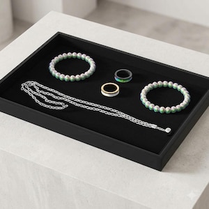 May include: A collection of jewelry items arranged on a black rectangular tray, including two beaded bracelets with iridescent green and silver spheres, a silver chain necklace, a black ring with a green inlay, and a gold-toned ring with a white band.