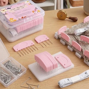 May include: A knit blocking combs set featuring a clear plastic storage box with a floral label, several pink plastic combs with metal pins, a small container of silver T-pins, and a white measuring tape with black markings in inches.