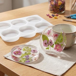 May include: A white silicone mold with six circular cavities, two clear circular resin coasters embedded with pressed pink flowers and green leaves, and a ceramic mug on a light-colored fabric cloth, with craft supplies in the background.