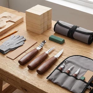 May include: A wood carving kit featuring three knives with dark wooden handles and metal blades, a gray fabric tool roll, a pair of gray cut-resistant gloves, a yellow pencil, a rectangular sharpening stone, a leather strop, and several wooden blocks.