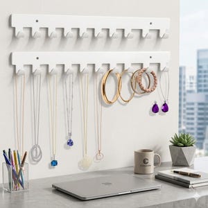 May include: Two white wall-mounted jewelry organizers with hooks, displaying necklaces, earrings, and bracelets. Gold and silver jewelry hangs from the hooks. A clear pen holder, a laptop, and a mug are on the desk.