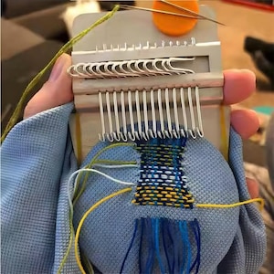 May include: A close-up of a loom with a small woven piece of fabric in blue, yellow, and white. The fabric is attached to a round, blue cushion. The loom has a metal frame with rows of hooks.