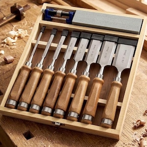 May include: A set of eight wood chisels with wooden handles and metal blades labeled CR-V, arranged in a wooden storage box alongside a sharpening stone and a blue honing guide. The blades vary in width, with the largest clearly marked as 50mm.
