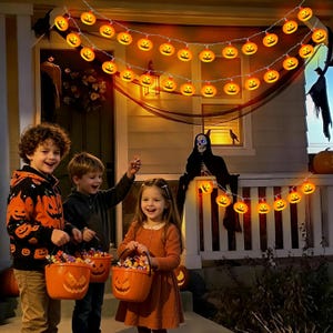 May include: A string of orange pumpkin-shaped LED lights hanging above three children holding orange plastic jack-o'-lantern buckets filled with candy. The scene features a skeleton decoration on a porch railing and dark fabric draped overhead.
