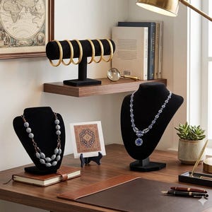 May include: A display of jewelry on a wooden desk. Gold bracelets are on a black stand. Two necklaces are displayed on black busts. A small rug is on a stand. A magnifying glass and books are on a shelf.