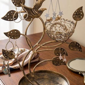 May include: A bronze-colored metal jewelry organizer shaped like a tree with a bird, leaf-shaped earring holders with small holes, a wire mesh basket for accessories, and a circular base tray for rings and small items, featuring a distressed finish.
