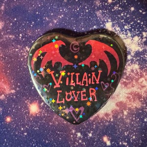 K&ouml;nnte beinhalten: Herzf&ouml;rmiger Button mit schwarzem Hintergrund und den Worten "Villain Lover" in Pink. Der Button zeigt rote Fledermausfl&uuml;gel und bunte Sternakzente. Der Hintergrund ist ein Galaxiedruck.