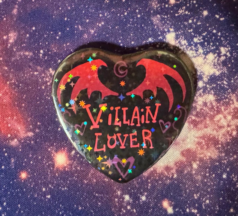 K&ouml;nnte beinhalten: Herzf&ouml;rmiger Button mit schwarzem Hintergrund und den Worten "Villain Lover" in Pink. Der Button zeigt rote Fledermausfl&uuml;gel und bunte Sternakzente. Der Hintergrund ist ein Galaxiedruck.