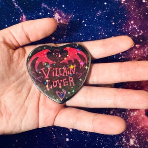 K&ouml;nnte beinhalten: Ein herzf&ouml;rmiger Button mit den Worten "Villain Lover" in rosa Schrift. Der Button hat einen schwarzen Hintergrund mit roten Fledermausfl&uuml;geln und Sternen. Der Button wird in einer offenen Hand vor einem Galaxiehintergrund gehalten.