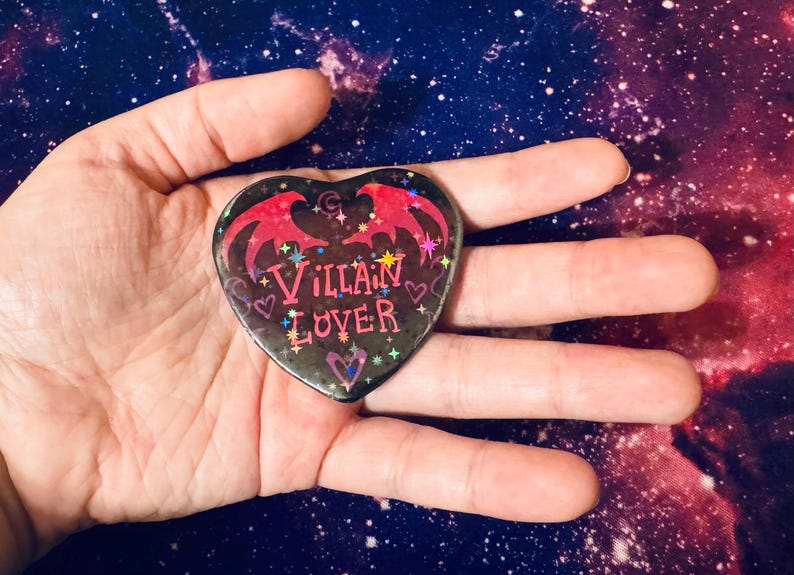 K&ouml;nnte beinhalten: Ein herzf&ouml;rmiger Button mit den Worten "Villain Lover" in rosa Schrift. Der Button hat einen schwarzen Hintergrund mit roten Fledermausfl&uuml;geln und Sternen. Der Button wird in einer offenen Hand vor einem Galaxiehintergrund gehalten.