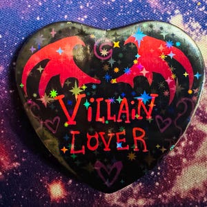 K&ouml;nnte beinhalten: Herzf&ouml;rmiger Button mit den Worten "Villain Lover" in Rot. Der Button hat einen schwarzen Hintergrund mit roten fledermausartigen Fl&uuml;geln und bunten Sternakzenten. Ein einzigartiges Accessoire.