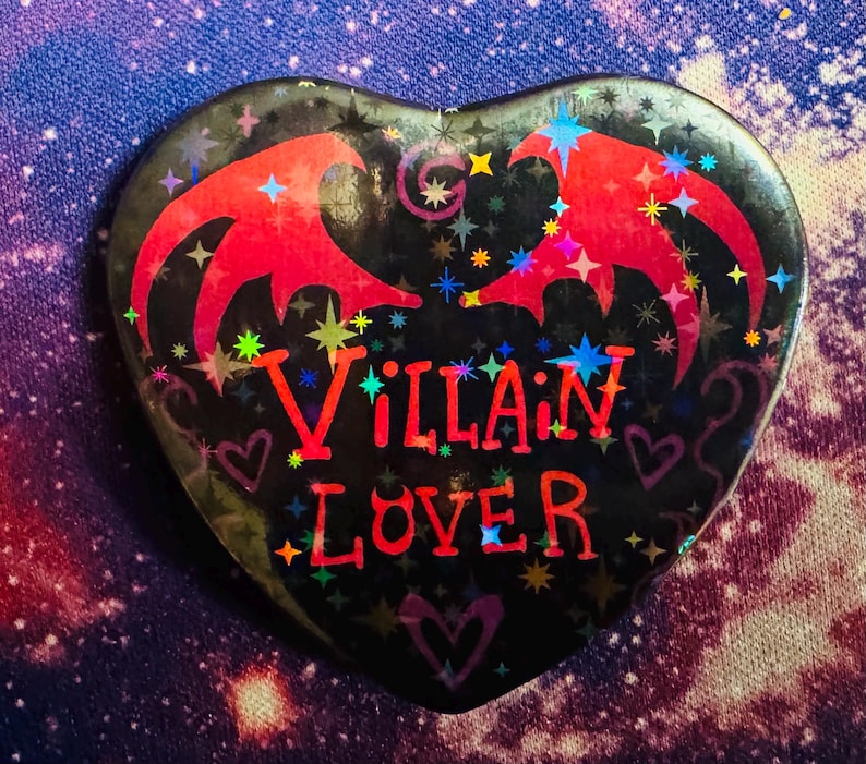 K&ouml;nnte beinhalten: Herzf&ouml;rmiger Button mit den Worten "Villain Lover" in Rot. Der Button hat einen schwarzen Hintergrund mit roten fledermausartigen Fl&uuml;geln und bunten Sternakzenten. Ein einzigartiges Accessoire.