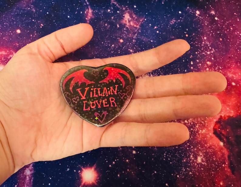K&ouml;nnte beinhalten: Herzf&ouml;rmige Anstecknadel mit den Worten "Villain Lover" in Rot und Pink auf schwarzem Hintergrund. Die Anstecknadel hat rote Fledermausfl&uuml;gel und dekorative Wirbel. Die Anstecknadel wird in einer Hand vor einem Hintergrund im Galaxien-Design gehalten.