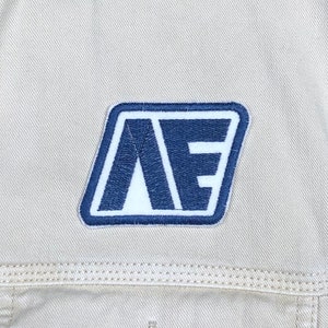 May include: Close-up of a tan denim jacket with a navy blue and white patch. The patch features the letters "AE" in a stylized font, outlined in white and set against a navy blue background.