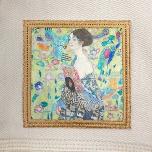 May include: A framed painting of a woman with a fan in a garden setting. The woman is wearing a blue and white dress and has long dark hair. The garden is filled with flowers and birds.