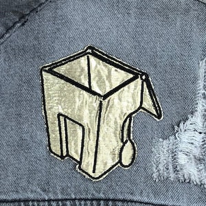 May include: Close-up of a gray denim jacket with a distressed patch. The patch is a gold-colored, stylized graduation cap design with black outlines. The jacket has a distressed area near the patch.