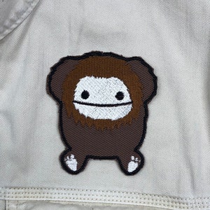 May include: A brown and white embroidered patch of a cartoon creature with a brown beard and white eyes. The creature has a brown body and white feet.