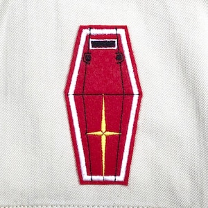 May include: A red and white embroidered patch with a yellow star design. The patch is in the shape of a shield and is sewn onto a beige fabric. The patch has a black border and a black rectangle at the top.