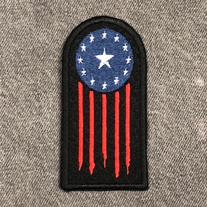 May include: A black and red embroidered patch with a circle of 13 stars and a single star in the center, representing the original 13 colonies of the United States. The patch has a distressed look with red stripes resembling a flag.
