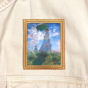 May include: A beige denim jacket with a rectangular patch featuring a painting of a woman holding a green umbrella. The painting has a gold frame and depicts a woman and child in a field under a blue sky.