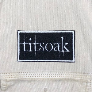 May include: Black and white embroidered patch with the text "titsoak" on a beige fabric background.