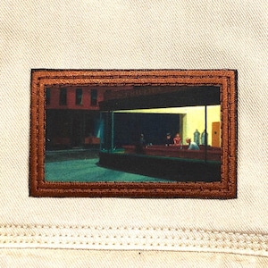 May include: A brown leather patch with a stitched border featuring a painting of a diner at night. The diner has a large window with people sitting at the counter. The painting is in a muted color palette of blues, greens, and browns.