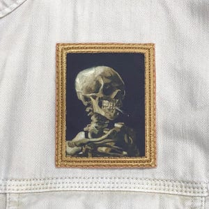 May include: A gold-framed embroidered patch featuring a detailed image of a human skull with a cigarette in its mouth. The skull is depicted in a realistic style, with visible bone structure and a dark background.