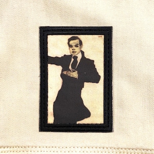 May include: A beige denim jacket with a rectangular patch featuring a sepia-toned illustration of a person in a suit and tie. The patch has a black border and is sewn onto the jacket.