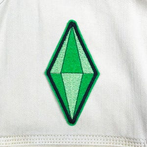 May include: A green diamond-shaped patch with a black border, reminiscent of a video game icon, is centered on a light-colored fabric. The patch features varying shades of green, creating a textured appearance. The fabric has visible stitching.