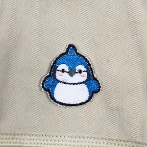 May include: A blue and white embroidered penguin patch with a white belly and black eyes. The penguin is smiling with pink cheeks.