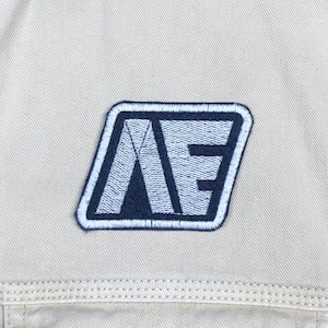 May include: A white patch with a blue and white embroidered logo of the letters "AE" in a geometric design.