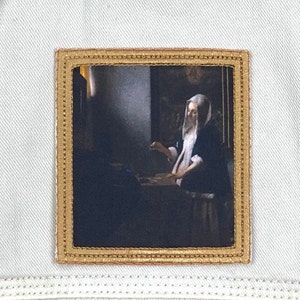 May include: A framed painting of a woman in a white head covering, sitting at a table in a dimly lit room. The painting is in a gold frame with a decorative border.