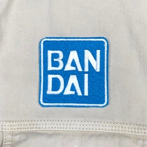 May include: A blue square patch with white stitching around the edges. The patch has the word "BANDAI" written in white letters.