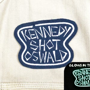 May include: A navy blue and white patch with the text "KENNEDY SHOT OSWALD" in a stylized font. The patch is on a light beige fabric. A smaller image shows the patch glowing in the dark.