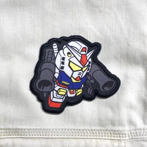 May include: A fabric patch featuring a stylized, chibi-style robot character with a white, blue, red, and yellow color scheme. The robot has a large head, yellow visor, and is holding two large black cannons on its shoulders, all outlined in black.
