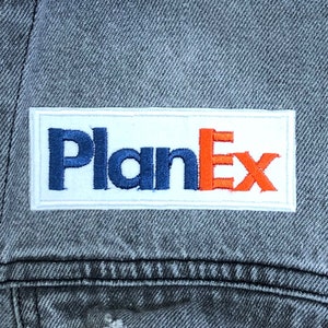 May include: A white rectangular patch with the text "PlanEx" in blue and orange lettering sewn onto a gray denim jacket.