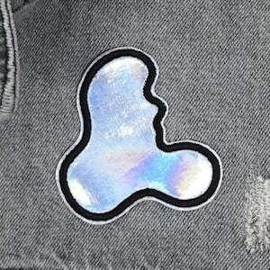 May include: A silver iridescent patch in the shape of a blob with a black outline. The patch is sewn onto a gray denim jacket.