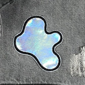 May include: A silver iridescent patch in the shape of a cloud with a black outline sewn onto a gray denim fabric.
