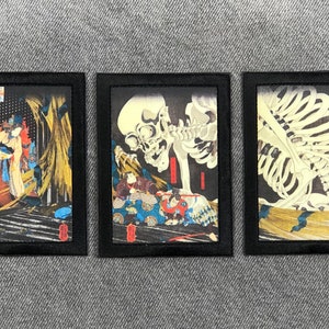 May include: Three framed black and white prints of Japanese woodblock prints depicting skeletons and figures in traditional clothing. The prints are set against a dark background.