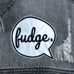 May include: A white speech bubble patch with black stitching and the word "fudge." sewn onto a gray denim jacket.