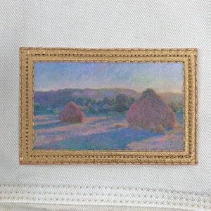 May include: A framed painting of a landscape with two haystacks in the foreground. The sky is a soft blue and purple, and the haystacks are a muted pink and brown. The painting is in a gold frame.