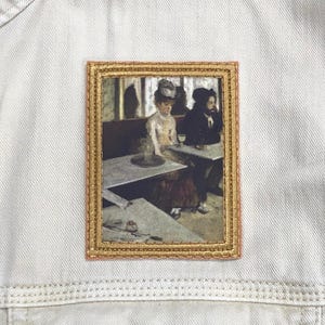 May include: A framed painting of two people sitting at a table in a cafe. The woman is wearing a hat and a dress, and the man is wearing a suit. The painting is in a gold frame.