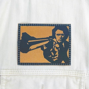 May include: A cream-colored denim jacket with a rectangular patch featuring a black and orange illustration of a person playing a trumpet. The patch is bordered with a dark blue stitched design.