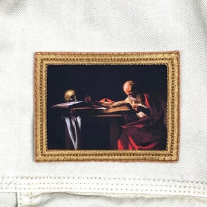 May include: A framed painting of a man with a long white beard, wearing a red robe, sitting at a table and writing in a book. A skull is on the table in front of him.