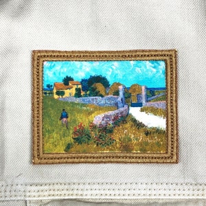 May include: A framed embroidered patch depicting a rural landscape with a yellow house, a stone wall, and a path leading to a gate. The sky is blue with white clouds, and there is a figure walking in the distance.