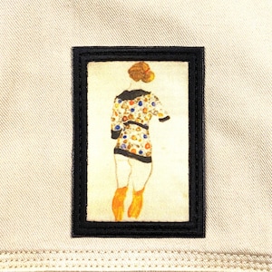 May include: A rectangular patch with a black border features a watercolor illustration of a person from the back, wearing a patterned top and orange boots. The patch is set against a beige fabric background.