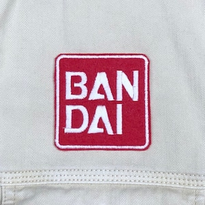 May include: Red square patch with white embroidered text that reads "BANDAI".